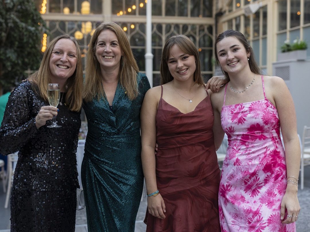 Four women standing closely together, smiling, and dressed in elegant evening gowns. One woman holds a champagne glass, and the setting features warm lighting and glass architecture in the background.